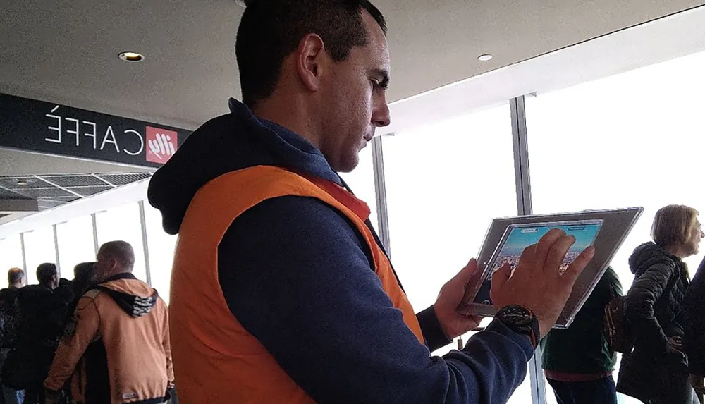 A man in a high-visibility vest is focusing on a tablet while standing in an indoor space with other people around