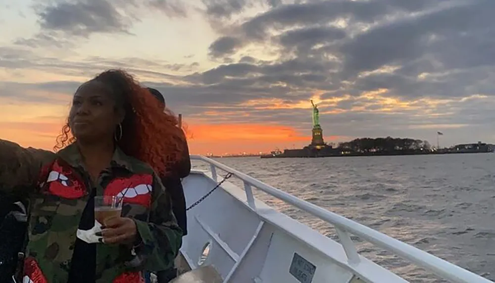 A person enjoys a drink on a boat with a view of the Statue of Liberty against a sunset sky