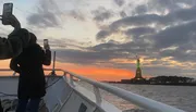 Two people are taking photos with their smartphones of the Statue of Liberty against a beautiful sunset sky from a boat.