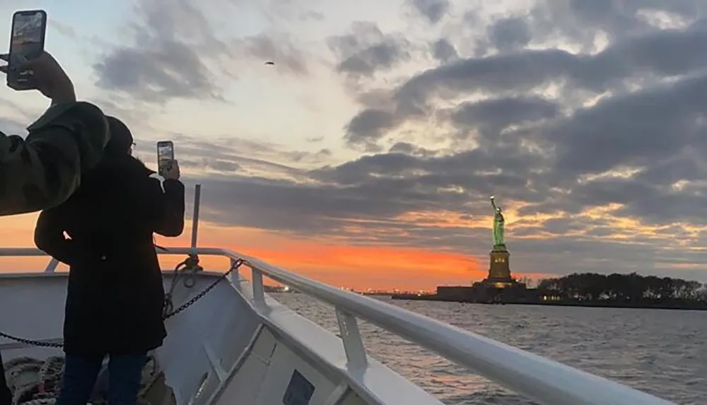 Two people are taking photos with their smartphones of the Statue of Liberty against a beautiful sunset sky from a boat