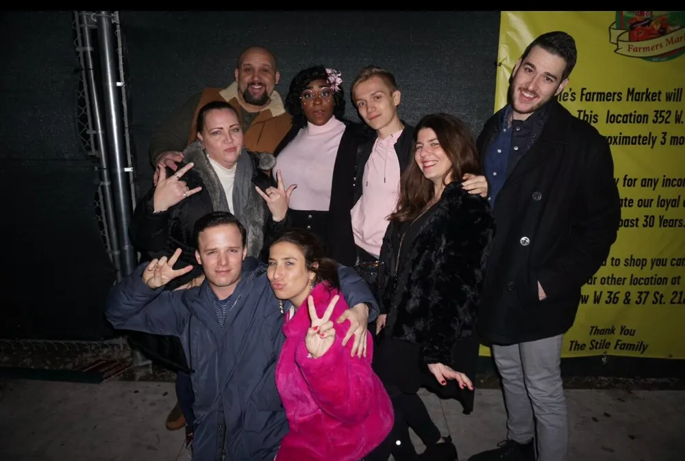 A diverse group of eight people is posing for a photo with some making peace signs smiling and enjoying what appears to be a casual night out