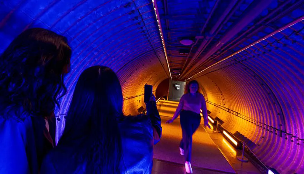 Two people are observing a third person walking towards them in an illuminated arched tunnel with blue and purple lighting