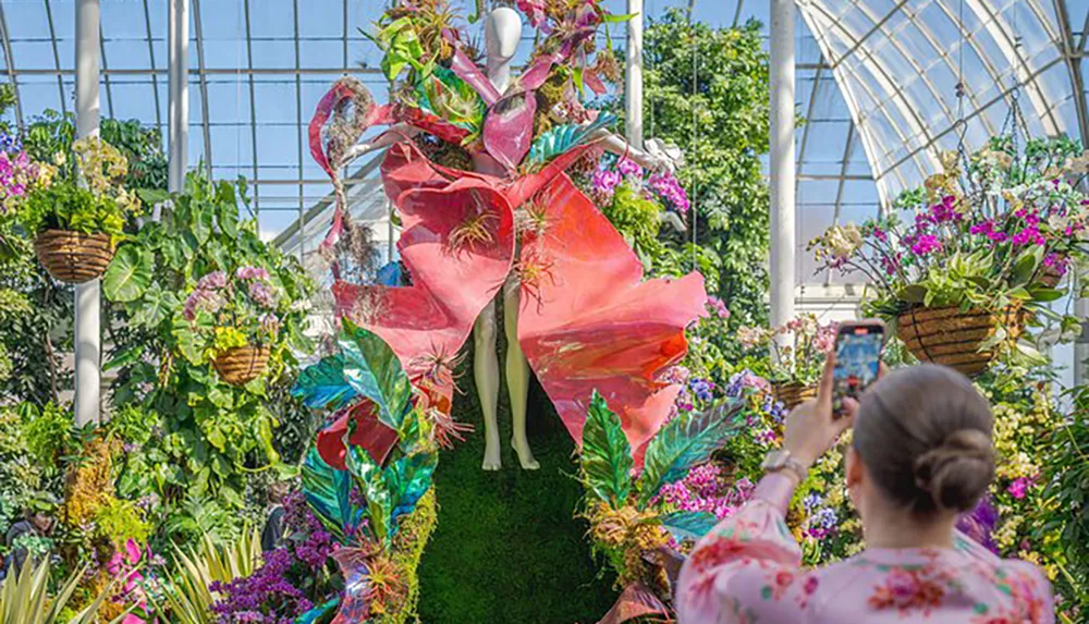 A person is photographing a vibrant botanical display featuring a mannequin dressed in large flower-like attire inside a sunlit greenhouse