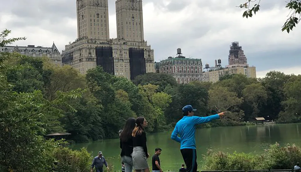 A person in a blue jacket is pointing at something in the distance while standing beside a lake in a park with tall buildings in the background likely in an urban setting