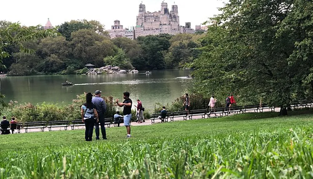 People enjoy a leisurely day near a pond in a lush urban park with a backdrop of high-rise buildings
