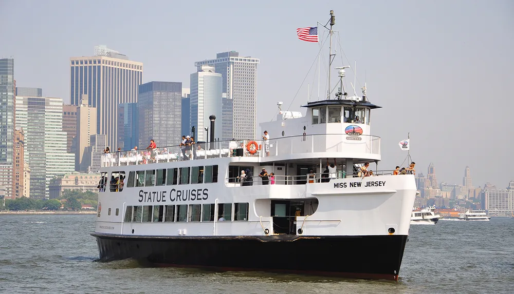 A ferry labeled Miss New Jersey from Statue Cruises is navigating the waters with passengers on board with the New York City skyline in the background