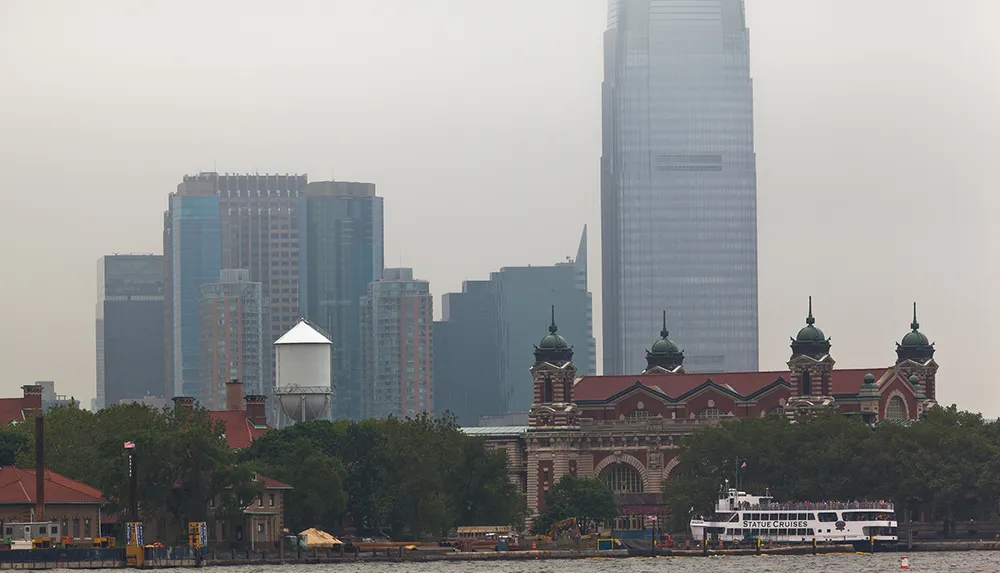 The image shows a contrast between an older ornate building in the foreground and the modern skyscrapers shrouded in haze in the background with a ferry labeled Statue Cruises moving along the water in front of the building