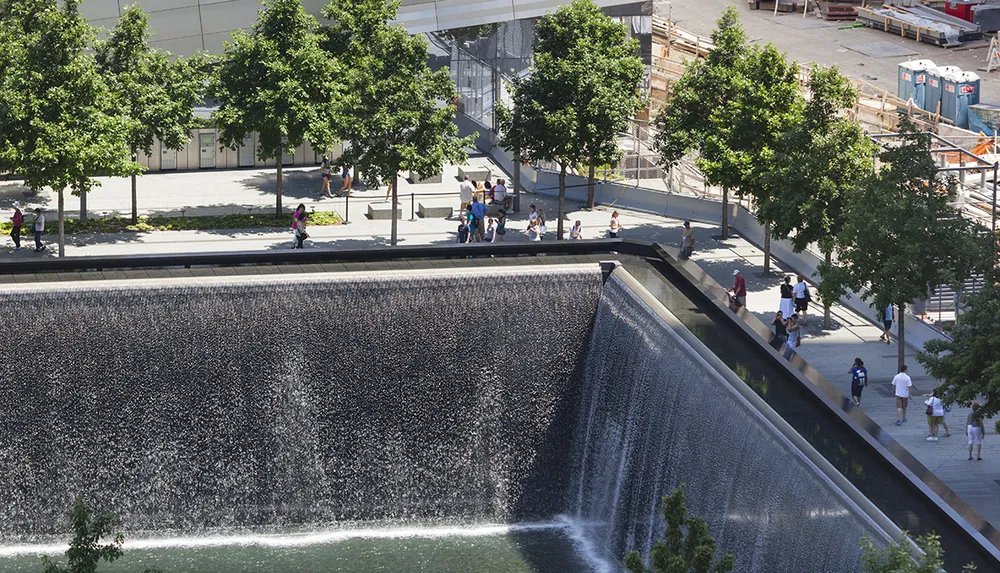The image shows a large cascading water feature in an urban park setting with people walking and enjoying the area on a sunny day