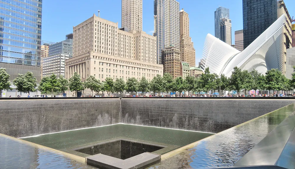 The image shows the 911 Memorial reflecting pools in Lower Manhattan with the Oculus structure in the background on a clear sunny day