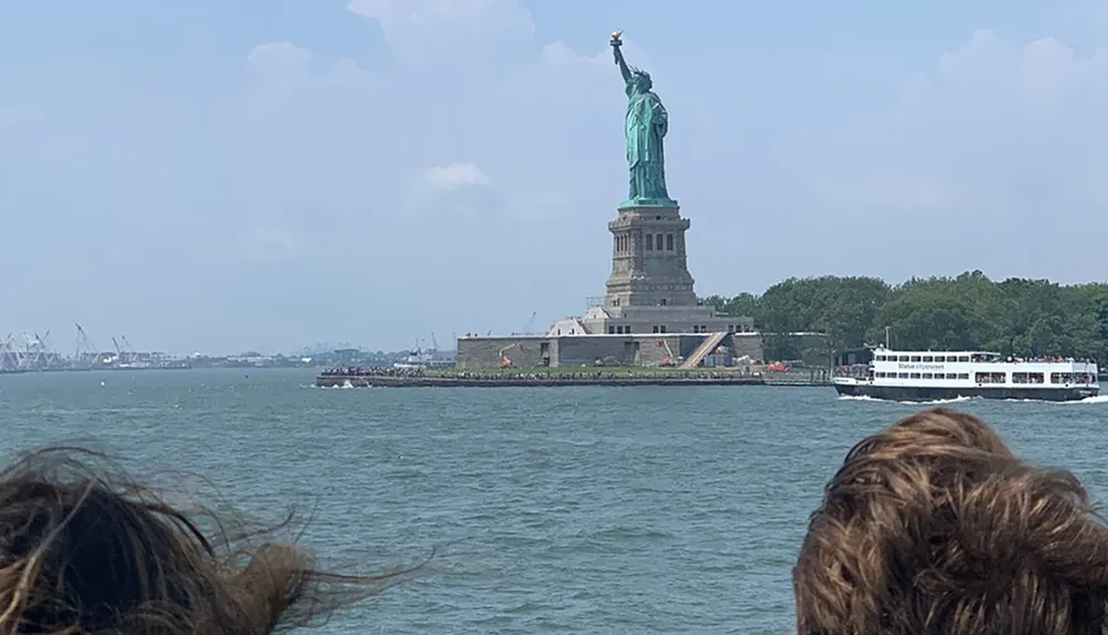 The image captures the iconic Statue of Liberty standing tall on Liberty Island as viewed from the water with people in the foreground and a ferry passing by