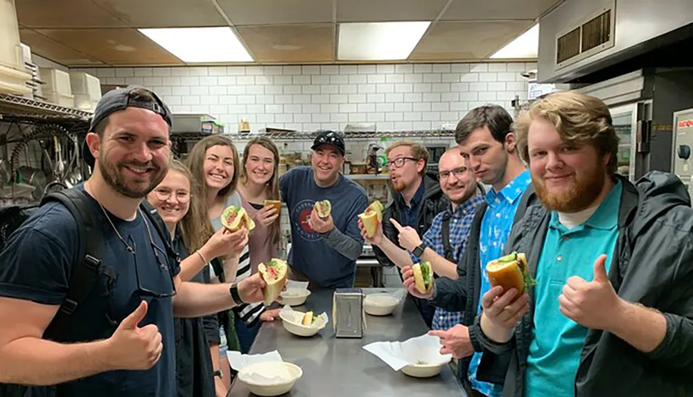 A group of people is cheerfully posing in a kitchen holding up sandwiches and giving thumbs-up signs