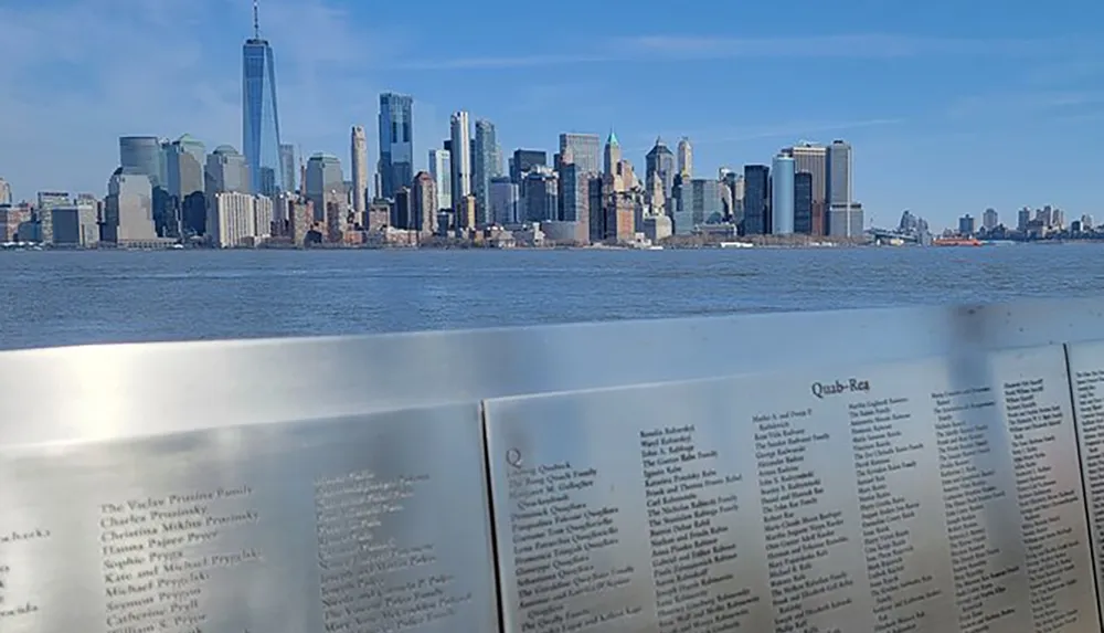 The image shows a view of the Manhattan skyline across a body of water seen over a reflective memorial panel with engraved names